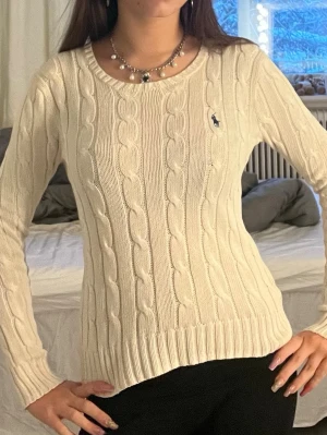 Vit kabelstickad tröja Polo Ralph Lauren - Kabelstickad vit tröja från Polo uppe med klassisk marinblå logga på bröstet. Tillverkad i mjuk pima bomull och har rund halsringning samt ribbade muddar. Perfekt för lager på lager och ger en clean vibe.