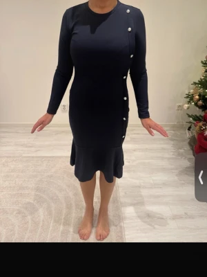 Marinblå figurnära klänning med knappar - Snygg marinblå klänning i figurnära modell med långa ärmar och dekorativa vita knappar längs ena sidan. Klänningen har rund halsringning och volangkant nedtill som ger en extra touch. Perfekt för dig som gillar stilrena detaljer och en feminin siluett.