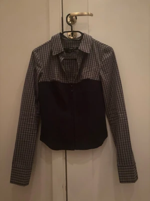 Rutig skjorta från Urban outfitters - Unik långärmad skjorta från Urban outfitters med rutigt mönster i grått och svart upptill och enfärgad svart nedtill. Skjortan har klassisk krage och dragkedja framtill för en modern twist. Perfekt för dig som vill sticka ut lite extra. Inte använt en enda gång pga tyvärr för liten