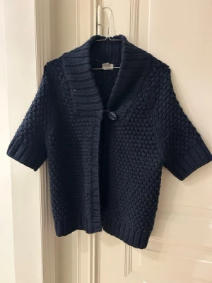 Mörkblå stickad kofta från H&M - Säljer en mörkblå stickad kofta från H&M med korta ärmar och grovstickad struktur. Koftan har en bred krage och stängs med en stor knapp framtill. Perfekt att slänga över en t-shirt för en chill look.