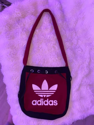Röd axelväska från Adidas - Snygg röd axelväska/påse från Adidas med svart kant och stort vitt logotryck framtill. Väskan har snörning upptill med metallöljetter och bred axelrem. Tillverkad i bomullstyg och har en enkel, sportig look som passar perfekt till streetwear. Mått: höjd, 29 cm ca. Bredd: ca 30 cm. 