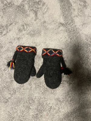 Svarta ullvantar med färgglada detaljer - Mörkgråsvarta ullvantar med broderade detaljer i rött, gult och blått runt handleden. Vantarna har tofsar i samma färger och är fodrade med Thinsulate för extra värme. Perfekta för kalla vinterdagar och har en traditionell look.