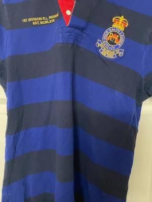Randig Ralph Lauren rugbytröja S - Säljer en blå och marinblå randig rugbytröja från Polo Ralph Lauren i custom fit. Tröjan har broderad logga på bröstet, gul textdetalj och klassisk krage med röd insida och vita knappar. Tillverkad i bomull och har korta ärmar.