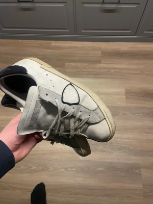 Vita Philippe Model sneakers i skinn - Säljer ett par vita Philippe Model sneakers med beige och svarta detaljer. Skorna har snörning, gummisula och en ikonisk sköld på sidan. Ovandelen är i skinn och mocka, med en klassisk låg profil och rund tå. Perfekta för dig som gillar stilrena och exklusiva sneakers.