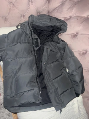 Grå pufferjacka från Canada Goose med scan och är jätte varm passar storlek M men även L  - Säljer en Grå pufferjacka från Canada Goose med luva och diskret logga på ärmen. Jackan har dragkedja och tryckknappar framtill, stora fickor och är perfekt för kalla dagar. Materialet är slitstarkt och jackan är riktigt varm och skön.