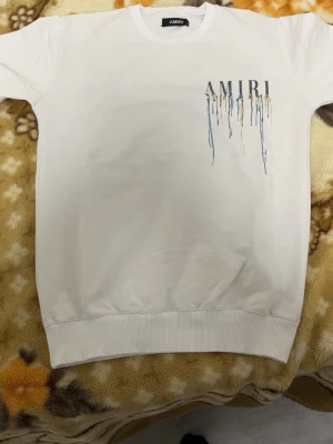 Vit sweatshirt från Amiri med färgstänk - Säljer en vit sweatshirt från Amiri med coolt färgstänksmönster i blått, gult och svart både fram och bak. Stor AMIRI-logga på ryggen och mindre logga på bröstet. Tröjan har långa ärmar och ribbade muddar vid ärmslut och nederkant. Perfekt för dig som gillar streetwear vibes.