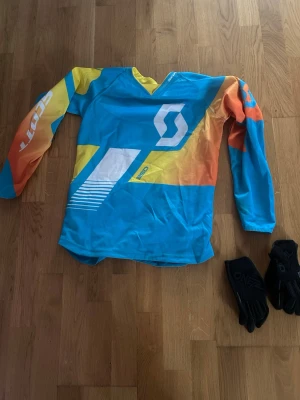 kross tröja - Bilden visar en färgglad långärmad tröja från Scott i blått, gult och orange samt ett par svarta handskar 