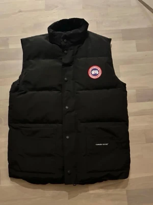 Svart dunväst från Canada Goose - Säljer en riktig svart dunväst från Canada Goose med hög krage och tryckknappar framtill. Västen har två stora fickor och den klassiska Canada Goose-loggan på bröstet. Perfekt för lager-på-lager och riktigt snygg till höst- och vinteroutfits.