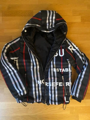 Jacka reversible  - Säljer en snygg från Burberry med klassiskt rutigt mönster i svart, vitt, grått och rött. Jackan är vändbar med ena sidan helsvart och andra sidan med stora Burberry-tryck. Utrustad med huva, dragkedja och justerbar nederkant.