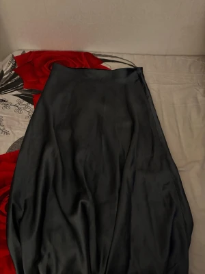 Svart långkjol i satin - Säljer en svart långkjol i glansigt satinliknande material. Kjolen har en enkel och stilren design med lätt A-linjeformad siluett som ger ett snyggt fall. Perfekt att styla med en crop top eller stickad tröja för en trendig look.