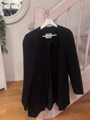 Svart kappa från Vero Moda - Stilren svart kappa från Vero Moda med rak passform och rundad halsringning. Jackan har diskreta fickor på sidorna och är helfodrad för extra komfort. Perfekt för dig som gillar en clean och enkel look.