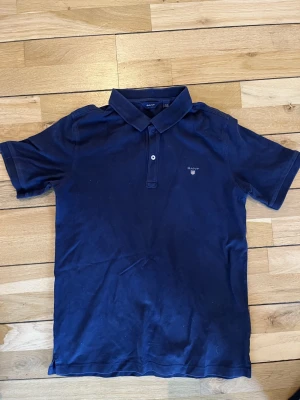 Marinblå pikétröja från Gant - Klassisk marinblå pikétröja från Gant med korta ärmar och krage. Tröjan har två knappar framtill och en liten broderad Gant-logga på bröstet. Tillverkad i mjuk bomull som känns skön mot huden.