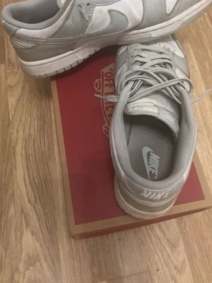 Nike Dunk låga sneakers i grått och vitt - Säljer ett par Nike Dunk sneakers i lågt utförande med snygg färgkombination av ljusgrått och vitt. Skorna har klassisk snörning, rund tå och är tillverkade i syntet och textil. Perfekta för dig som gillar stilrena och tidlösa sneakers.