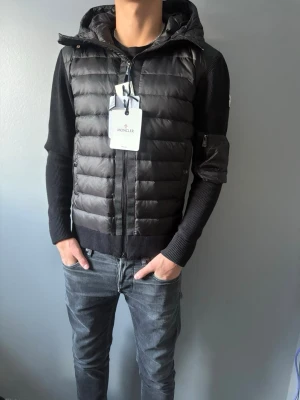 Svart Moncler cardigan jacka - Säljer en svart pufferjacka från Moncler med stickade ärmar och vadderad kropp. Jackan har huva, dragkedja framtill, fickor med knappstängning och en liten ficka med dragkedja på ärmen. Perfekt för dig som vill ha en stilren och modern look. Helt ny oanvänd. Scannen funkar vid tvättlappen. NFC taggen funkar också vid moncler märket.