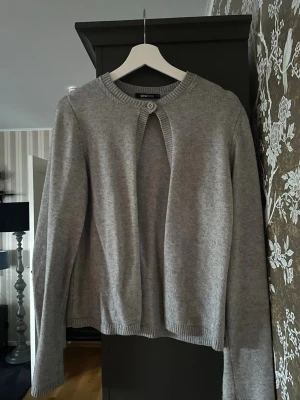 Grå kofta från Gina Tricot - Mysig grå kofta från Gina Tricot i stickad bomullsblandning. Klassisk rund halsringning med ribbade kanter och en knapp framtill som ger en enkel men snygg look. Perfekt att slänga över en topp under kyligare dagar.