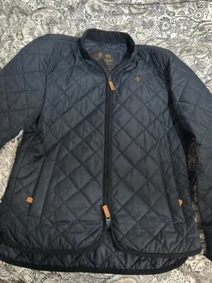 Quiltad mörkblå jacka från Morris - Säljer en quiltad mörkblå jacka från Morris i storlek small. Jackan har dragkedja framtill, två fickor med snygga läderdetaljer och ett diskret broderat emblem på bröstet. Perfekt för dig som gillar stilren och klassisk design. Jackan är i väldigt bra skick men höger ficka har ett hål men inget man ser utan msn känner det bara! Nypris är 2499 kr och köpt för ett år sen och använd under höst och vår mitt pris är 1399! Kom med frågor. storlek S