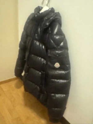 Svart Moncler pufferjacka - Säljer en svart pufferjacka från Moncler med glansig finish och klassisk logga på ärmen. Jackan har hög krage, dragkedja framtill och praktiska fickor. Perfekt för kalla dagar när du vill vara både varm och stilren.