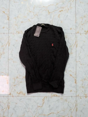 Svart kabelstickad tröja Polo Ralph Lauren - Kabelstickad svart tröja från Polo Ralph Lauren med rund halsringning och den klassiska röda logon broderad på bröstet. Tröjan har långa ärmar och ribbade muddar vid ärmslut och nederkant. Perfekt för dig som gillar stilren och tidlös design.