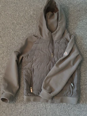 Grå pufferjacka med huva från Moncler - Säljer en beige pufferjacka från Moncler med huva och dragkedja framtill. Jackan har quiltade partier på bröstet och två fickor med dragkedja. Klassisk Moncler-logga på ärmen och ribbade muddar vid ärmslut och nederkant. Perfekt för kyliga dagar.