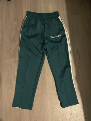 Gröna Palm Angels trackpants S - Mörkgröna trackpants från Palm Angels med vita sidoränder och logga på vänster ben. Byxorna har elastisk midja och raka ben. Materialet är 80% bomull och 20% polyester, vilket ger en mjuk och sportig känsla. Perfekta för en avslappnad streetwear-look.