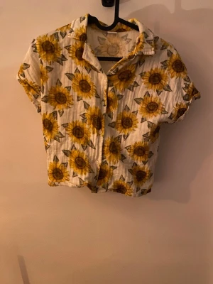 Solrosmönstrad skjorta från Lindex - Kortärmad skjorta från Lindex med stora solrosor i gult och grönt på vit botten. Skjortan har klassisk krage, knappar framtill och är gjord i mjuk bomull. Perfekt för dig som vill sticka ut med ett färgglatt och somrigt plagg.