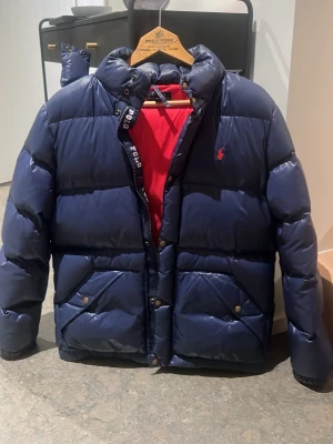 Mörkblå pufferjacka Polo Ralph Lauren XL - Säljer en mörkblå pufferjacka från Polo Ralph Lauren i storlek 170, passar folk mellan XS och S. Jackan har röd insida samt två stora fickor på framsidan. Klassisk Polo-logga broderad på bröstet och Polo-detaljer på dragkedjan🧥