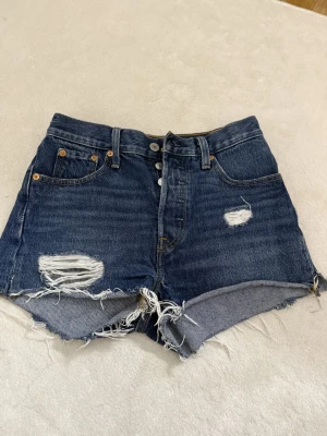 Levi's blåa high waist jeansshorts - Säljer ett par klassiska blåa jeansshorts från Levi's med hög midja och råa, fransiga kanter. Shortsen har slitna detaljer framtill, fem fickor och knäppning med knappar. Midjemått rakt över är 34cm!🫰