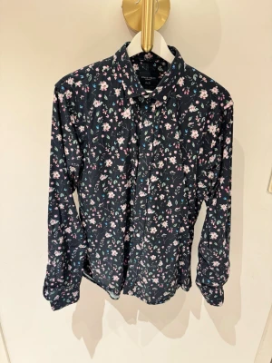 Blommig skjorta från Grand Frank - Snygg slim fit skjorta från Grand Frank i mörkblått med blommigt mönster i rosa, vitt och grönt. Skjortan har långa ärmar, klassisk krage och knäppning framtill. Perfekt för dig som vill sticka ut med en cool och färgglad look.