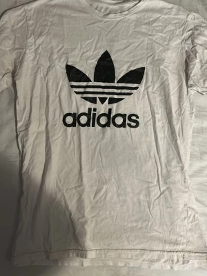 Vit Adidas t-shirt med svart logga - Vit t-shirt från Adidas med den klassiska svarta trefoil-loggan tryckt stort på bröstet. T-shirten har rund halsringning och korta ärmar. Tillverkad i mjuk bomull som är skön att bära hela dagen.