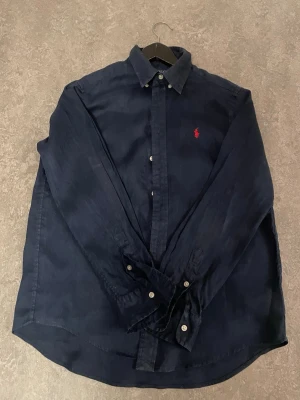Mörkblå skjorta från Polo Ralph Lauren - Mörkblå skjorta från Polo Ralph Lauren med klassisk krage och knappar framtill. Skjortan har det ikoniska röda broderade Polo-logot på bröstet och är tillverkad i mjuk bomull. Perfekt till en clean och stilren look. Själv är jag 1,67 och den passar mig utmärkt skulle säga att den passar dig som är runt 160-175 ungefär.kontakta mig för mer information och bilder om det behövs och självklart så tvättar jag den innan jag fraktar den