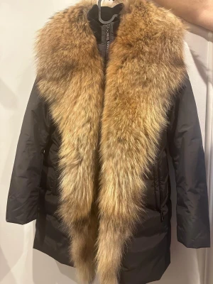 Svart dunjacka med stor päls från Hollies - Säljer en svart lång dunjacka från Hollies med en riktigt maffig, naturfärgad pälskrage. Jackan har dragkedja framtill, två fickor med dragkedja och en badge på ärmen. Perfekt för kalla vinterdagar när du vill vara både varm och snygg.