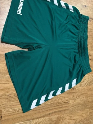 Gröna Hummel träningsshorts - Säljer ett par gröna träningsshorts från Hummel med vita detaljer längs sidorna och elastisk midja. Shortsen har en sportig look och är tillverkade i ett lätt och smidigt polyestermaterial, perfekta för träning eller sport.