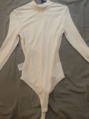 Vit långärmad polotopp body - Säljer en stilren vit body med lång ärm och hög krage. Toppen har en clean look utan mönster och knäppning nedtill. Perfekt att styla med jeans eller kjol för en enkel men snygg outfit.