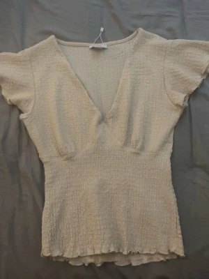 Beige ribbad topp med v-ringning - Säljer en beige ribbad topp med djup v-ringning och korta, lite fladdriga ärmar. Toppen har en smickrande passform och vågig kant längst ner. Perfekt till jeans eller kjol för en enkel men snygg look.