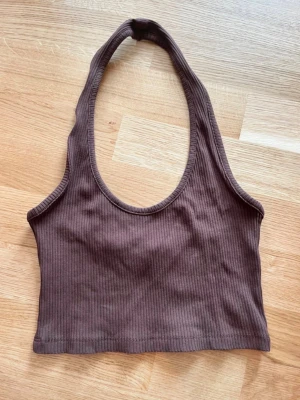 Brun ribbad croppad halternecktopp - Säljer en brun croppad topp med ribbad struktur och halterneck. Toppen är ärmlös och har en enkel, stilren design som passar perfekt till jeans eller kjol. Materialet känns mjukt och stretchigt, vilket gör den bekväm att bära.