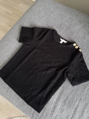 Svart kortärmad topp med guldknappar - Säljer en svart kortärmad topp från H&M i mjukt, strukturerat material. Toppen har rund halsringning och tre dekorativa guldknappar på ena axeln som ger en snygg detalj. Passar perfekt till både jeans och kjol för en clean look. Prislappen tog bort men tröjan kom aldrig till användning.