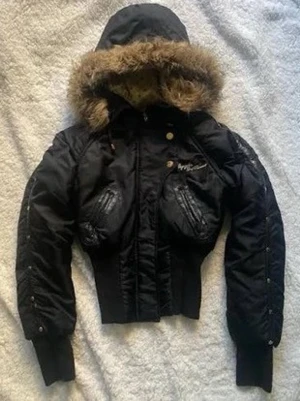 Svart bomberjacka från Apple Bottoms - Säljer en svart bomberjacka från Apple Bottoms med stor huva och fejkpäls runt kanten. Jackan har coola detaljer som guldknappar, ribbade muddar och stora fickor framtill. På ryggen finns en stor logga med nitar och broderi. Perfekt statement-plagg för dig som vill sticka ut.