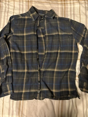 Rutig trekantsmönstrad skjorta Marmot - Säljer en mörkblå och beige rutig skjorta från Marmot i storlek S. Skjortan har långa ärmar, klassisk krage och knäppning framtill. Materialet är mjukt och skönt, perfekt för chill dagar. Detaljer som bröstficka och diskreta blå ränder i mönstret.