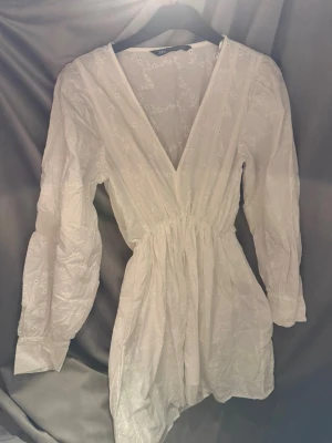 Vit broderad kortklänning från Zara - Snygg vit kortklänning från Zara med broderade blommor, v-ringning och långa ärmar. Klänningen har en markerad midja och lätt puffiga ärmar. 