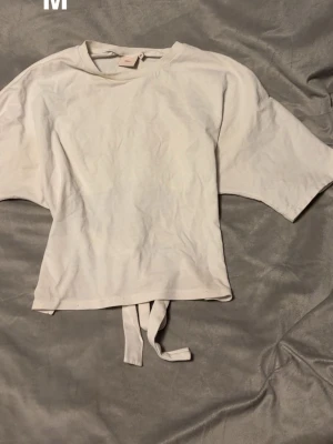 Vit croppad topp med knyt bak - Säljer en vit croppad topp från Nelly i mjuk bomull. Toppen har korta ärmar och ett snyggt knytband bak i midjan som ger en cool detalj. Enkel och clean design som passar till mycket.