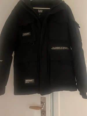 Svart pufferjacka från Black Squad - Säljer en svart pufferjacka från Black Squad med flera stora fickor och coola textdetaljer på bröstet och ärmen. Jackan har huva, dragkedja och en oversized look som ger streetwear-vibbar. Perfekt för kalla dagar och riktigt snygg till jeans eller cargo.