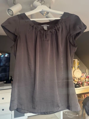 Mörkgrå sidenblus med kort ärm - Säljer en elegant mörkgrå blus i siden med rundad halsringning och lätt rynkade detaljer upptill. Blusen har korta ärmar och ett mjukt, glansigt material som ger en lyxig känsla. Perfekt att styla med jeans eller kjol för en trendig look.