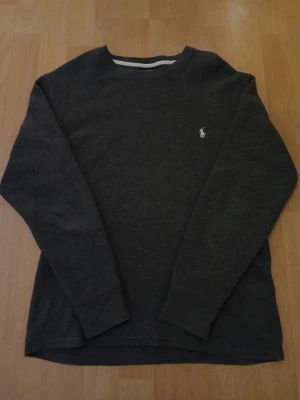 Mörkgrå stickad tröja från Polo Ralph Lauren. - Säljer en mörkgrå stickad tröja från Polo Ralph Lauren med klassisk rund hals och långärmad passform. Tröjan har en vit broderad logga på bröstet och är tillverkad i mjukt stickat material som är perfekt för kyliga dagar. Bredd: 48cm / Längd: 62cm. Nypris: 995kr / Mitt pris: 590kr