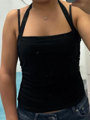 Svart halterneck linne med knyt - Säljer ett svart linne med halterneck och smala axelband. Toppen har en snygg knytning i nacken och är figurnära i modellen. Perfekt till jeans eller kjol för en enkel men stilren look.