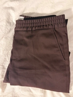 Bruna kostymbyxor från Filippa K - Snygga rödvina kostymbyxor från Filippa K med elastisk midja och raka ben. Byxorna har diskreta sidofickor och är gjorda i ett mjukt material som känns skönt mot huden. Perfekta för en clean och stilren look. Ull blandning 