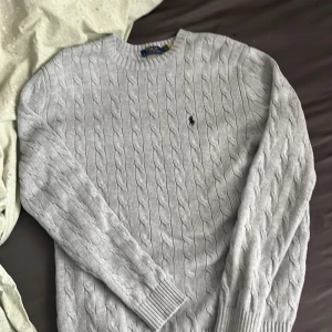 Ljusgrå kabelstickad tröja från Polo Ralph Lauren - Säljer en klassisk ljusgrå kabelstickad tröja från Polo Ralph Lauren med rund halsringning och broderad logga på bröstet. Tröjan har långa ärmar och ribbade muddar vid ärmslut och nederkant. Perfekt för lager-på-lager. Är storlek S men passar även M