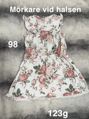 Blommig vit klänning med volang - Supersöt vit klänning från Newbie med rosa och grönt blommönster. Klänningen har volang vid halsen och spetsdetalj längst ner. Resår i midjan för skön passform och ärmlös design. Perfekt för varma dagar!🌸 Fläck vid halsen. Storlek 98