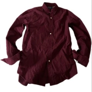Vinröd skjorta Polo Ralph Lauren - Snygg vinröd skjorta från Polo Ralph Lauren i slim fit-modell. Skjortan har klassisk krage, långa ärmar och knäppning framtill. Broderad grön logga på bröstet. Perfekt för dig som gillar stilrena och tidlösa plagg. Köptes för 1400 kr på Jonells. Säljer 499.