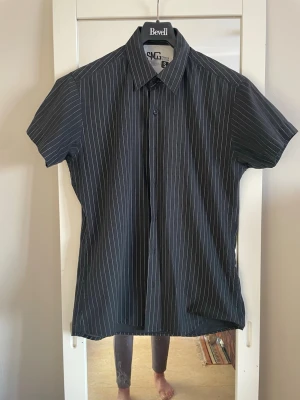 Svart randig skjorta från SMG - Kortärmad svart skjorta från SMG med tunna vita ränder och klassisk krage. Skjortan har knäppning framtill och en rak passform som ger en clean look. Perfekt till jeans eller chinos för en stilren vibe.