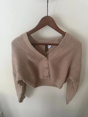 Beige croppad kofta från Gina Tricot - Säljer en beige croppad kofta från Gina Tricot med grovstickad struktur. Koftan har stora knappar framtill, v-ringad hals och långa ärmar. Perfekt för lager på lager och en chill look.